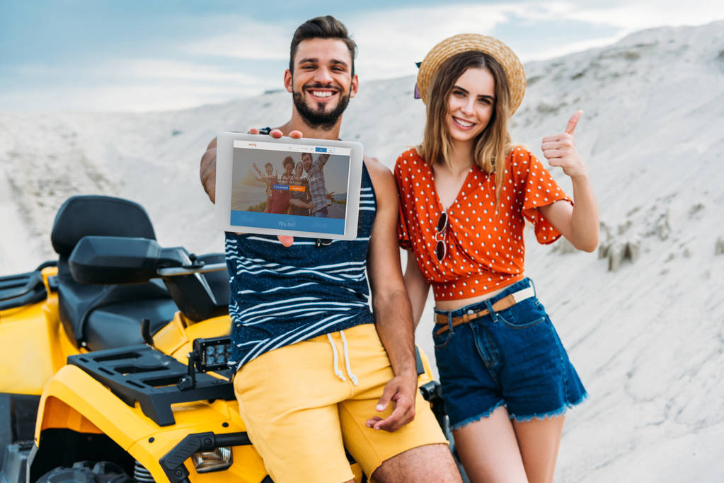 glückliches junges Paar mit ATV zeigt digitales Tablet mit Couchsurfing-Website auf dem Bildschirm und Daumen hoch in der Wüste - Foto, Bild