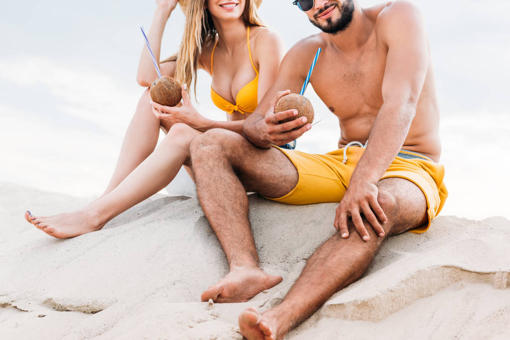 tiro recortado de pareja joven con cócteles de coco sentado en la arena frente al cielo nublado
 - Foto, imagen