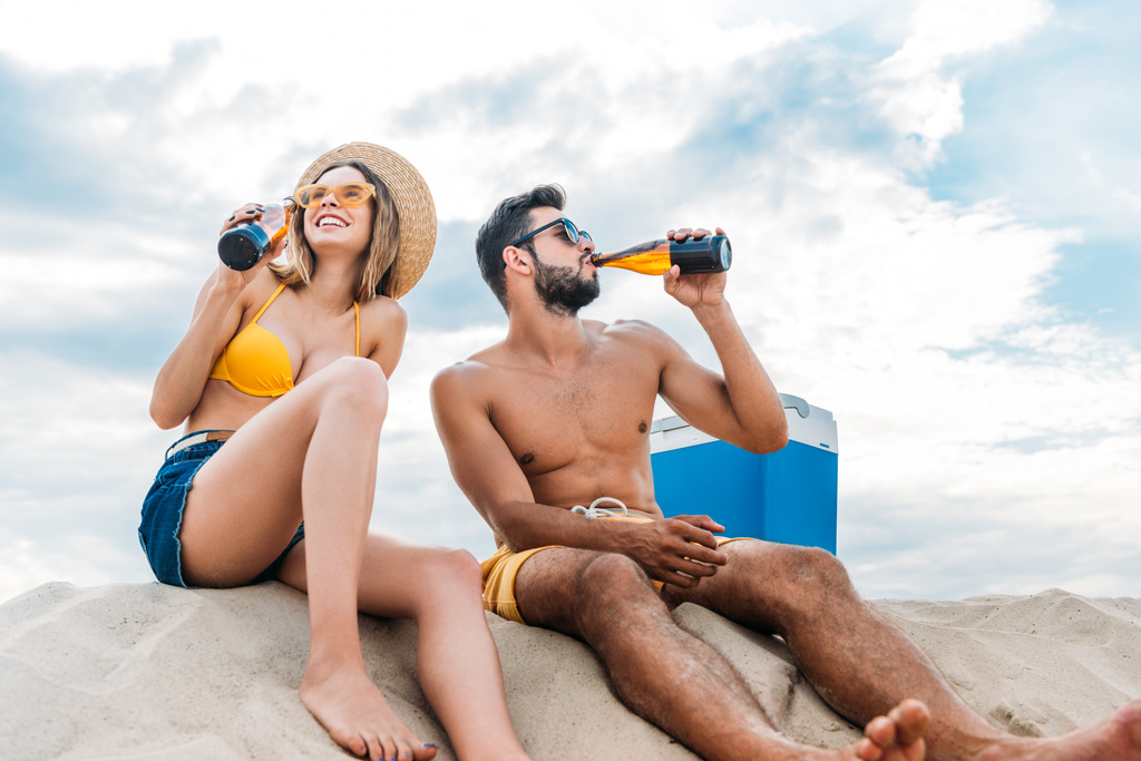 όμορφο νεαρό ζευγάρι πίνοντας μπύρα, ενώ κάθεται στην άμμο - Φωτογραφία, εικόνα