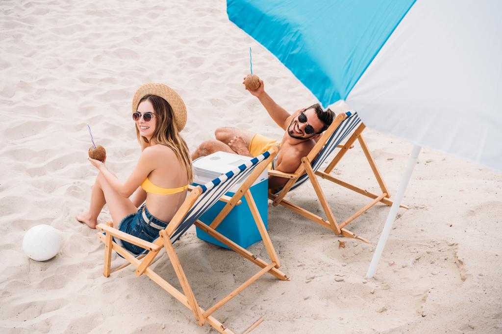 vista de alto ângulo de feliz jovem casal com coquetéis de coco relaxante em espreguiçadeiras na praia de areia
 - Foto, Imagem