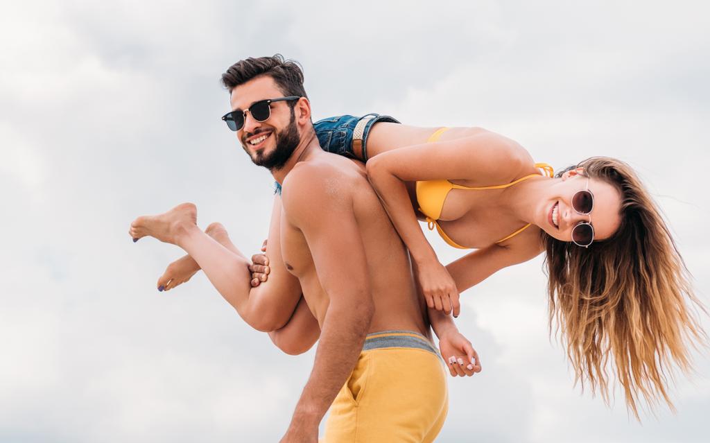 šťastný mladý muž nesoucí svou přítelkyni na rameni a při pohledu na fotoaparát - Fotografie, Obrázek