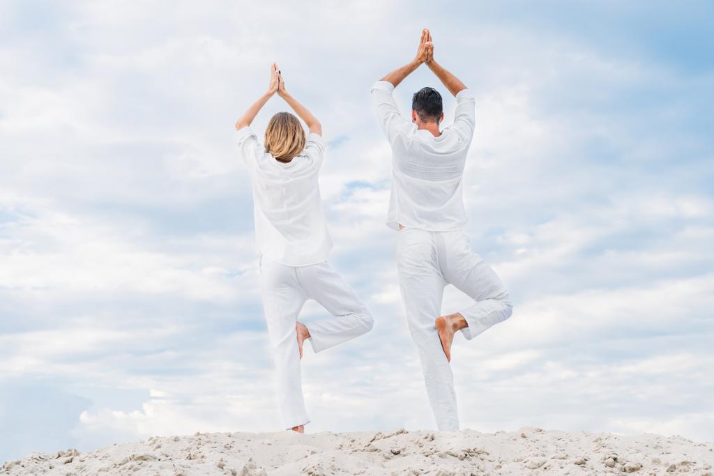 visão traseira do casal em roupas brancas praticando ioga em pose de árvore (Vrksasana) em duna arenosa
 - Foto, Imagem