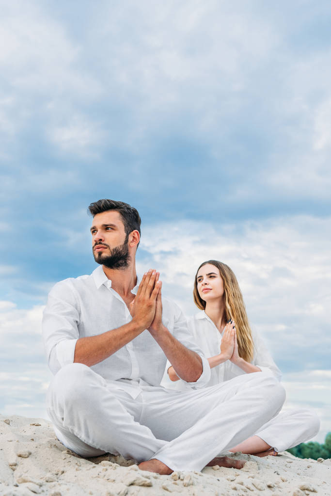 beautiful young couple practicing yoga while sitting on sandy dune in lotus pose (padmasana) and looking away - Photo, Image