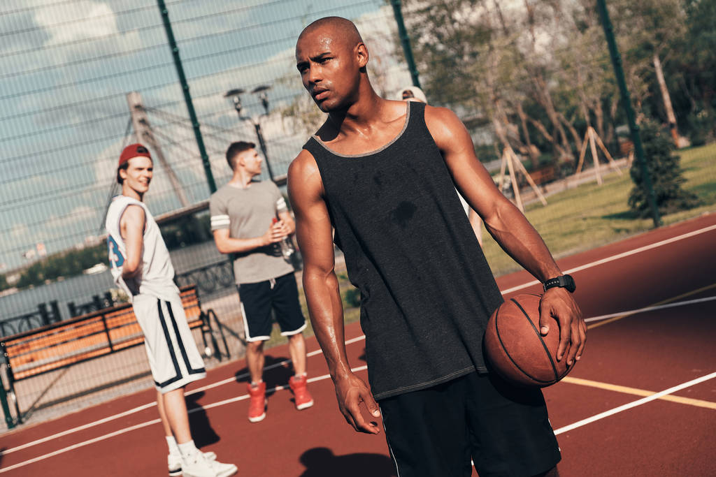 hombre africano deportivo mirando hacia otro lado mientras sostiene la pelota de baloncesto en la cancha con amigos
 - Foto, Imagen