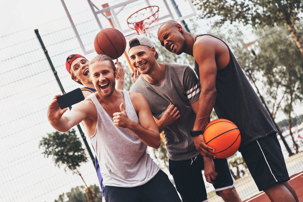 group of smiling men in sports clothing taking selfie on mobile phone while posing on basketball arena court - Photo, Image
