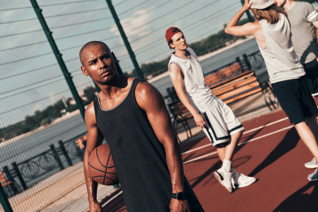 esportivo Africano homem olhando embora enquanto segurando bola de basquete na quadra com amigos
 - Foto, Imagem