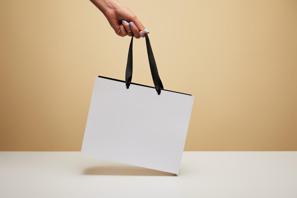 cropped image of woman holding white shopping bag above white table isolated on beige - Photo, Image