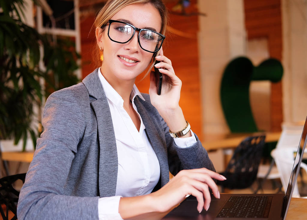 gute Geschäfte reden. junge schöne Frau mit Brille, die am Arbeitsplatz mit dem Handy telefoniert und mit dem Laptop lächelt. - Foto, Bild