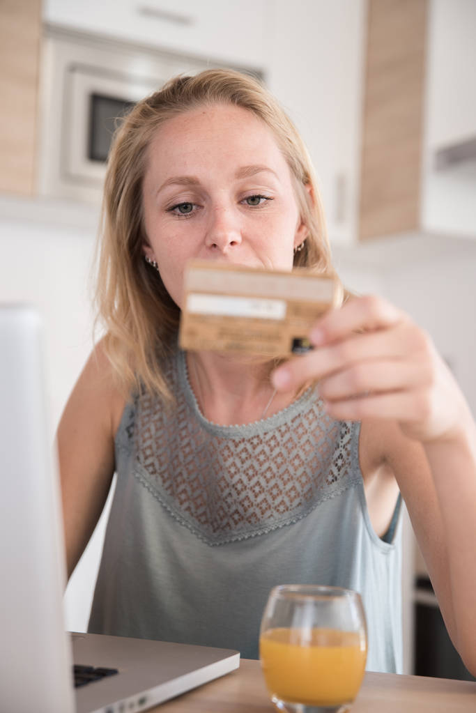Woman sitting at a table in a kitchen as she holds her credit card in her hand as she sits in front of a laptop - Photo, Image