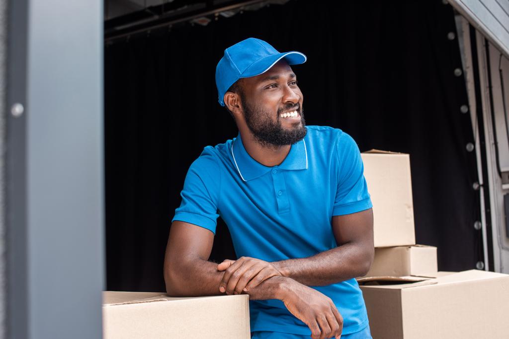 sonriente mensajero afroamericano apoyado en cajas y mirando hacia otro lado
 - Foto, imagen