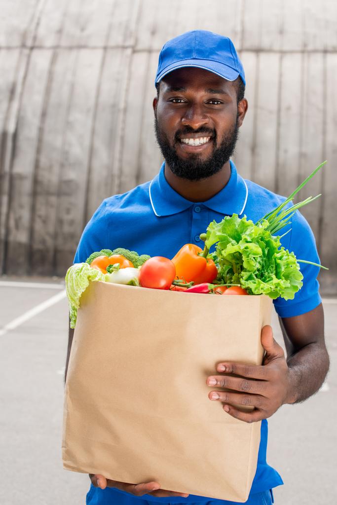 smiling african american delivery man holding paper bag with vegetables - Photo, Image