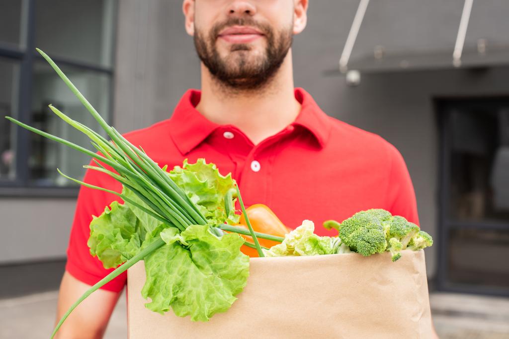 visão parcial do homem de entrega em saco de papel uniforme vermelho com legumes frescos
 - Foto, Imagem
