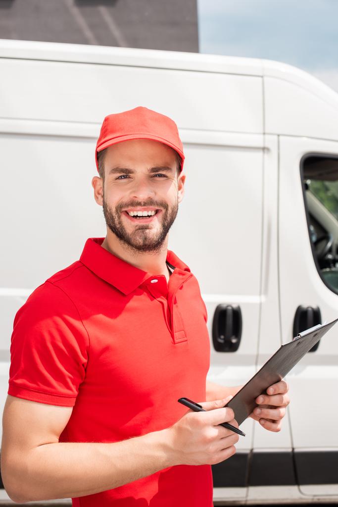 cheerful delivery man in red uniform with notepad looking at camera - Photo, Image