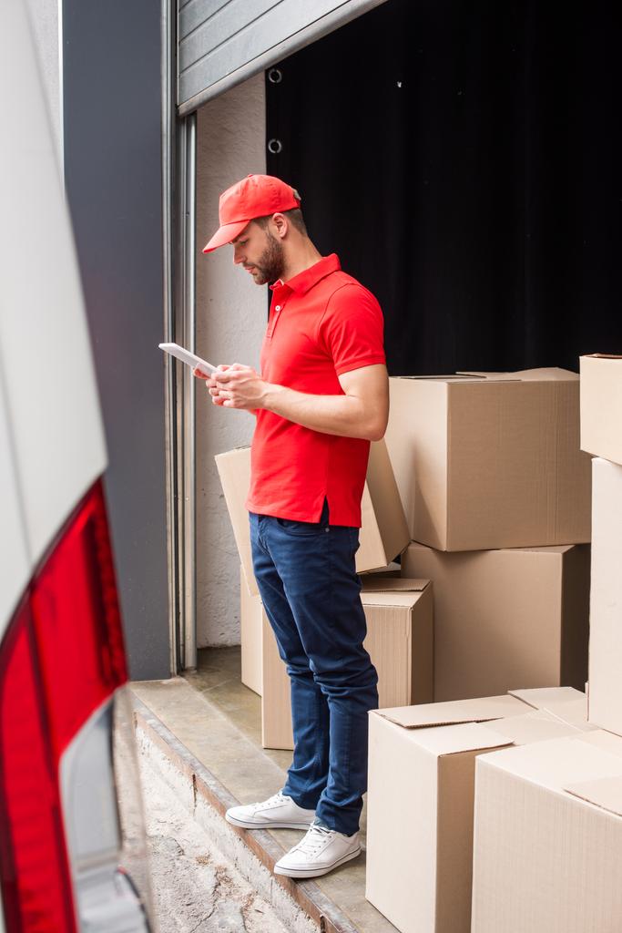 side view of delivery man in red uniform using digital tablet with cargo behind - Photo, Image