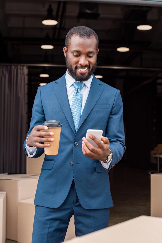 retrato de sonriente hombre de negocios afroamericano con café para llevar y smartphone
  - Foto, imagen