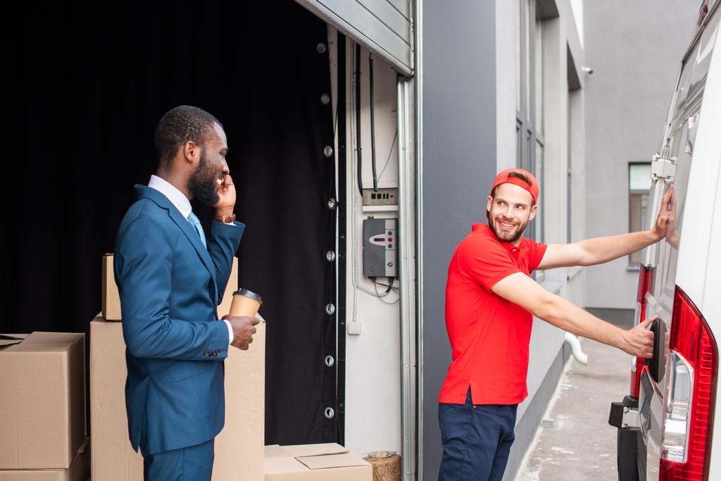 african american businessman with coffee to go controlling caucasian delivery man while talking on smartphone - Photo, Image