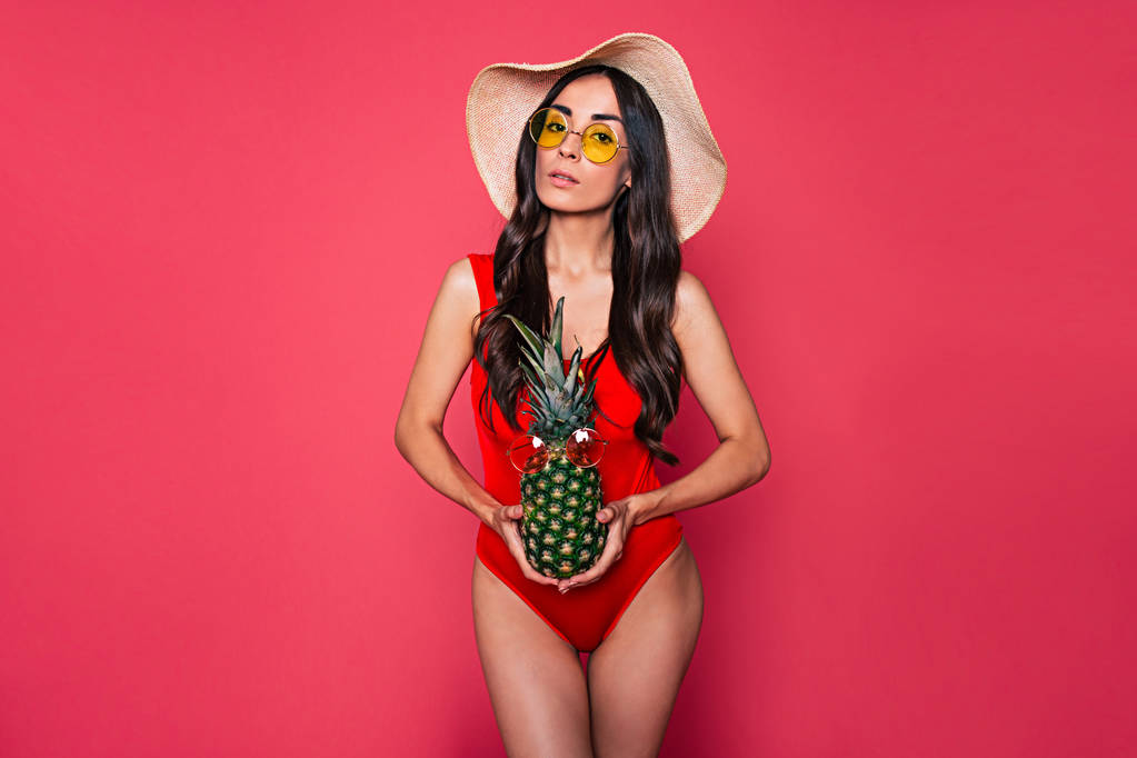 Genç esmer kadın kırmızı mayo, şapka ve gözlük pembe bir arka plan üzerinde ananas ile poz  - Fotoğraf, Görsel