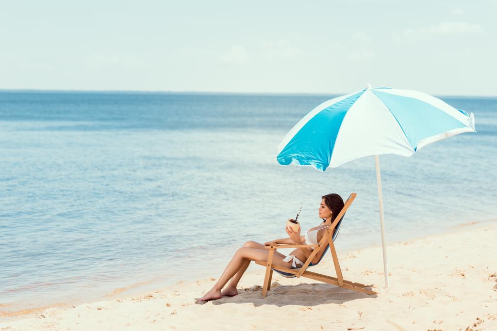beautiful girl in bikini with coconut cocktail resting on deck chair under beach umbrella near sea - Photo, Image