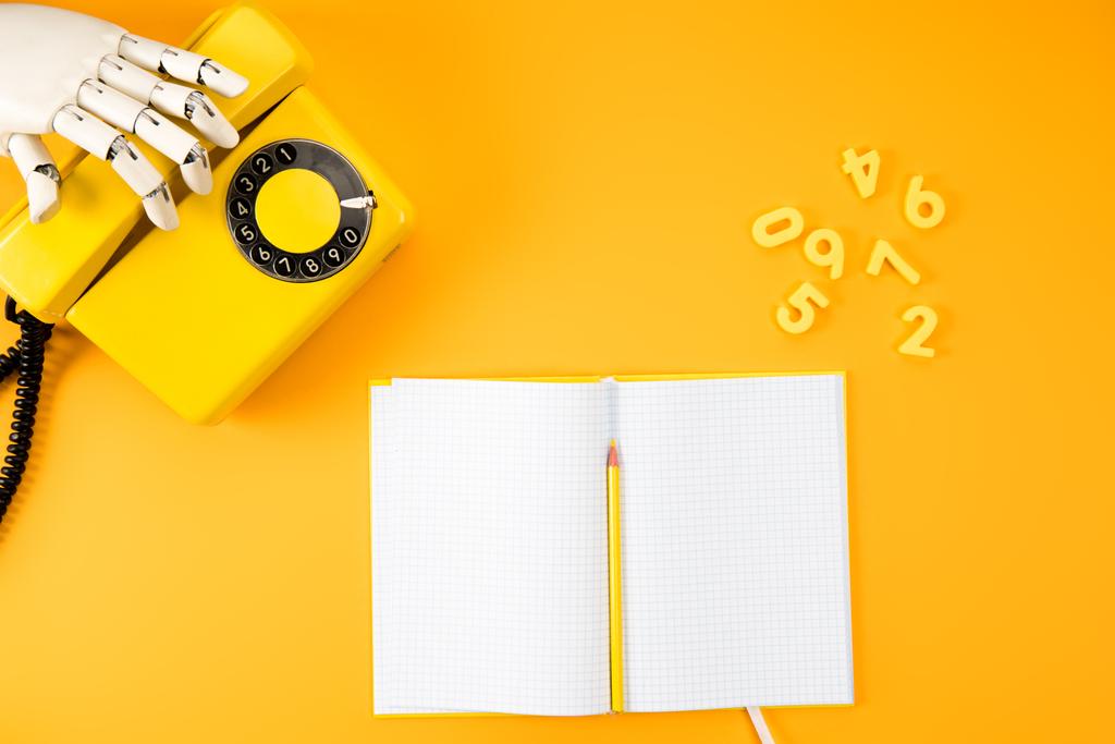 cropped shot of robotic hand reaching for vintage phone on yellow tabletop with blank notebook and math numbers - Photo, Image