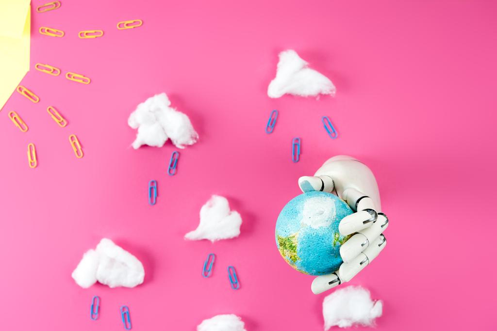 bovenaanzicht van regen samenstelling geregeld van paperclips en katoen met robotic hand met earth globe op roze - Foto, afbeelding