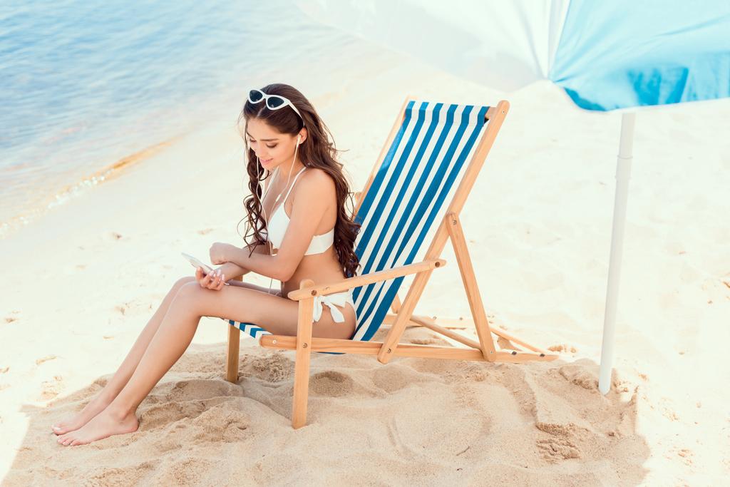 ελκυστική γυναίκα χρησιμοποιώντας smartphone και στηρίζεται σε παραλία καρέκλα κάτω από ομπρέλα κοντά στη θάλασσα στο θέρετρο - Φωτογραφία, εικόνα