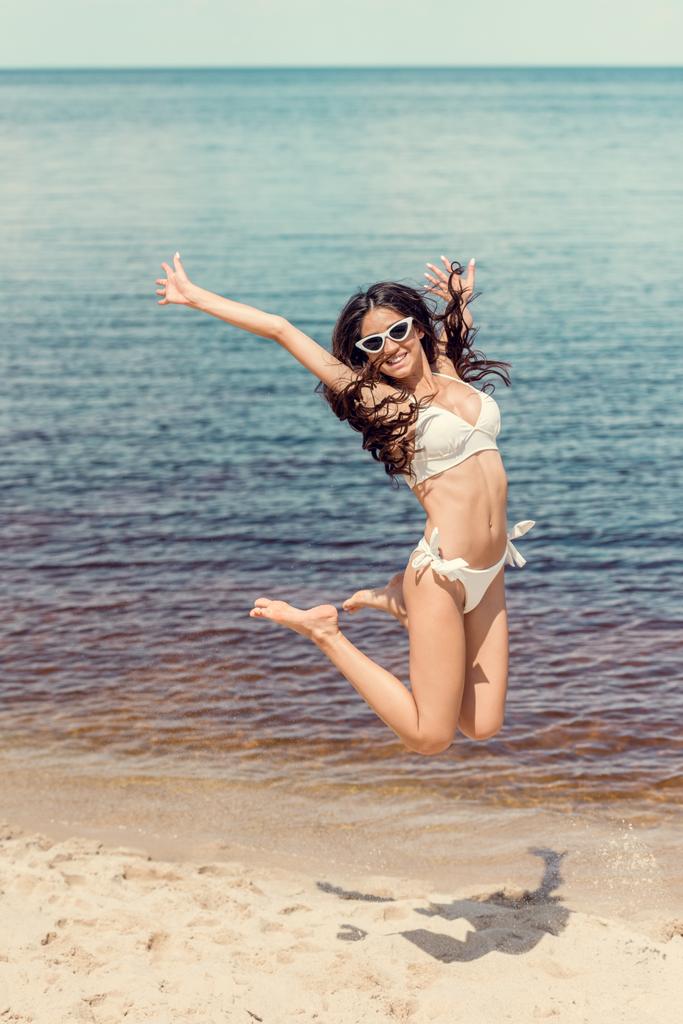 opgewonden vrouw in zonnebrillen en witte bikini op strand in de buurt van de zee springen  - Foto, afbeelding