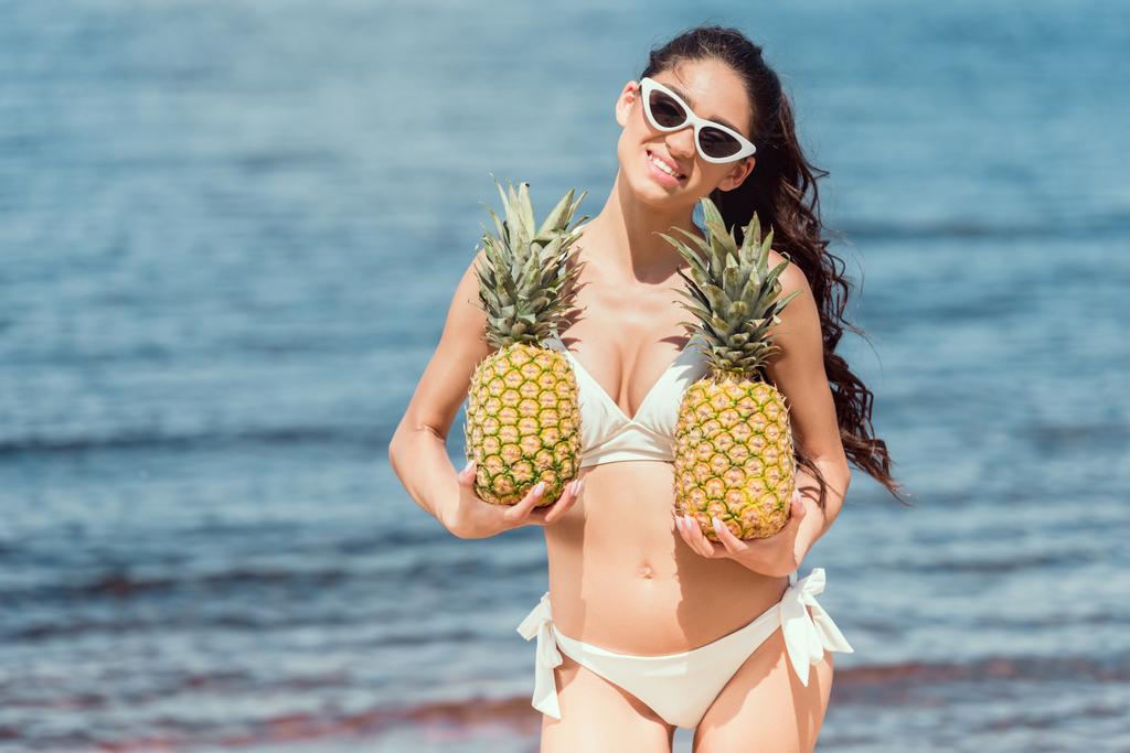 hermosa mujer sonriente en bikini y gafas de sol sosteniendo piñas frescas cerca del mar
 - Foto, imagen
