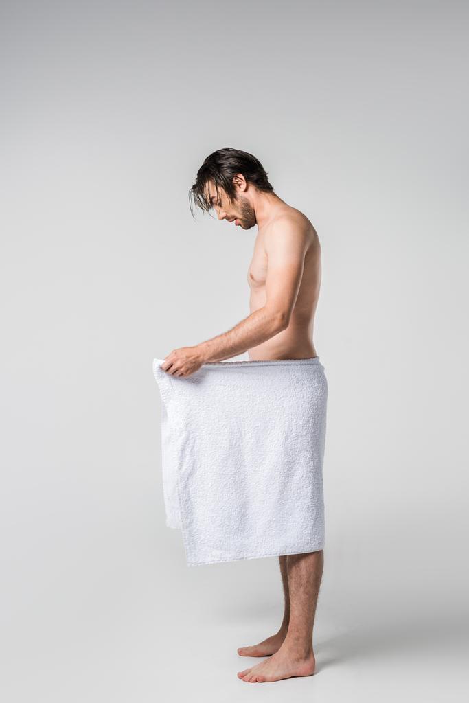 vista lateral de homem bonito em toalha branca olhando para baixo no fundo cinza
 - Foto, Imagem