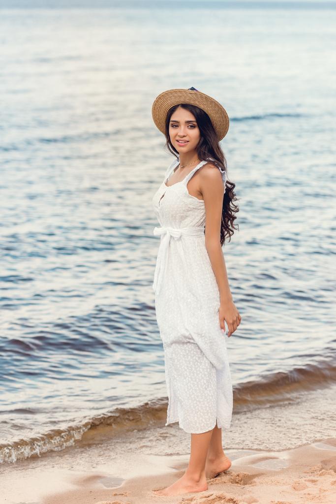 beautiful young woman in straw hat and white dress walking on sea shore - Photo, Image