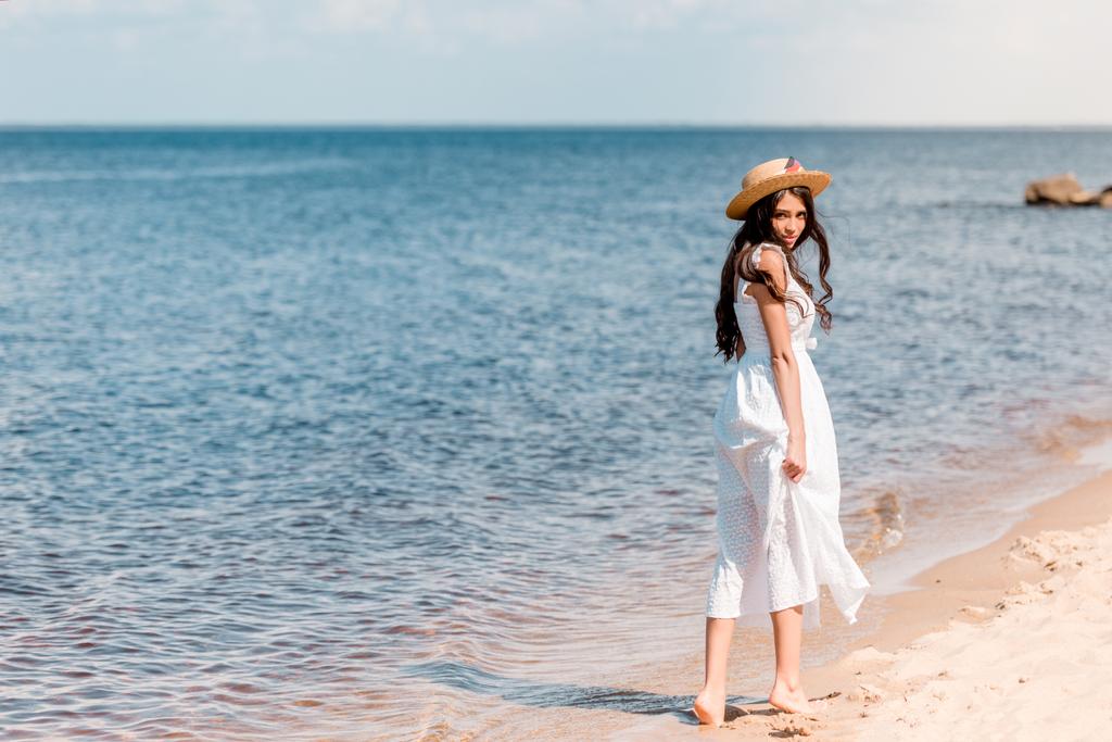 mujer joven en sombrero de paja y vestido blanco caminando en la playa
 - Foto, imagen