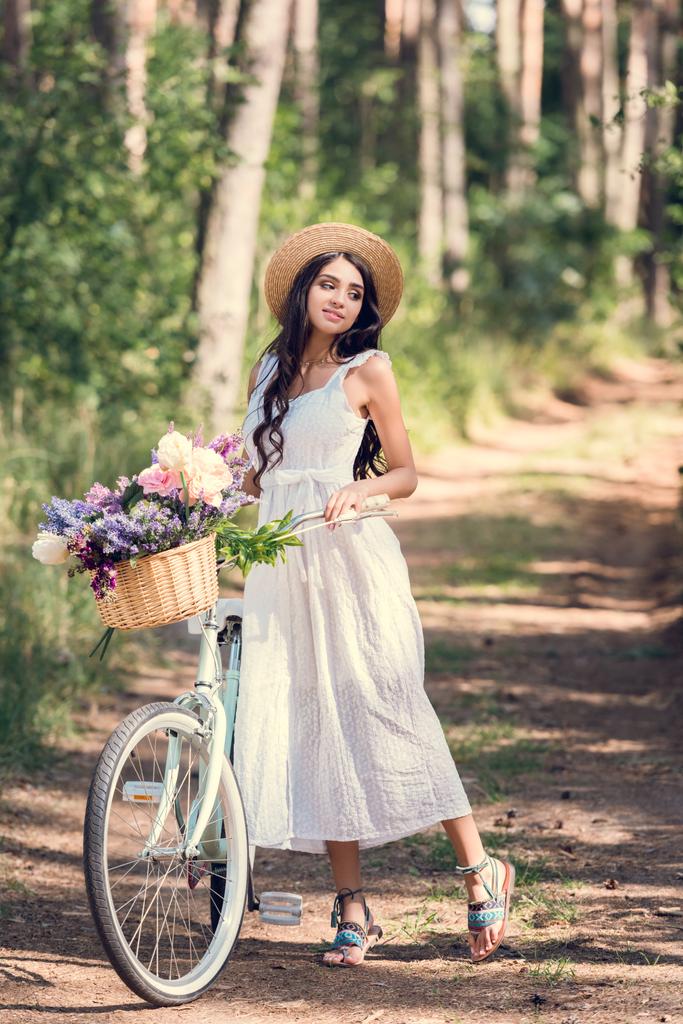ευτυχισμένος κορίτσι σε ψάθινο καπέλο και το λευκό φόρεμα που ποζάρει με ποδήλατο και ψάθινο καλάθι λουλούδια - Φωτογραφία, εικόνα
