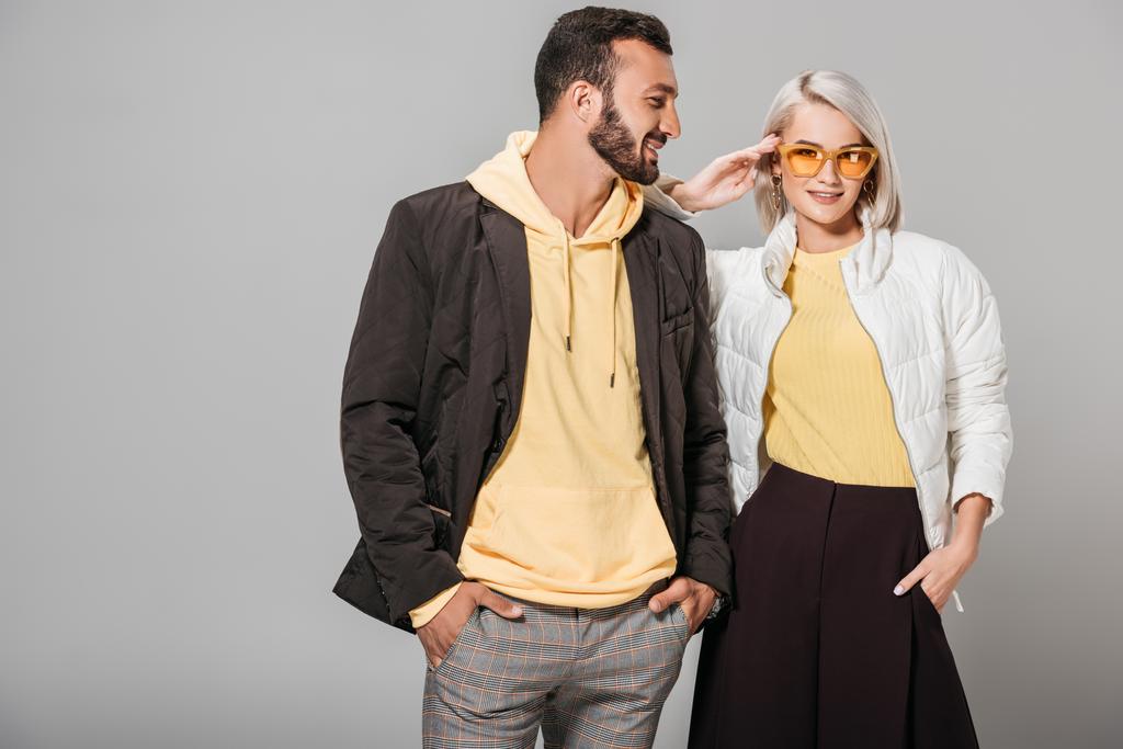 lachende mannelijk model met handen in de zakken poseren in de buurt van stijlvolle vriendin in zonnebril geïsoleerd op een grijze achtergrond  - Foto, afbeelding