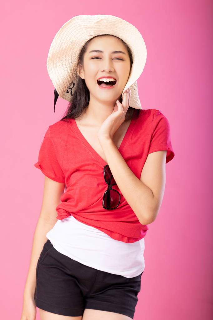 Πορτρέτο του ένα χαμογελαστό γυναίκα ελκυστική σε καλοκαιρινή στολή και καπέλο θέτοντας ενώ στέκεται και βλέπουν τα φωτογραφικών μηχανών που απομονώνονται σε ροζ φόντο. - Φωτογραφία, εικόνα