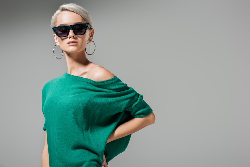魅力的な女性モデルのサングラスと灰色の背景上に分離されて腰に手でポーズ グリーンのセーター  - 写真・画像