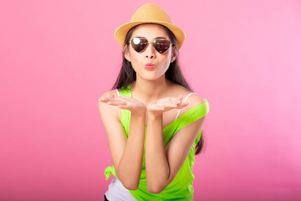 Retrato de una mujer atractiva sonriente en traje de verano verde y sombrero posando beso mientras está de pie y mirando a la cámara aislada sobre el fondo rosa
. - Foto, imagen