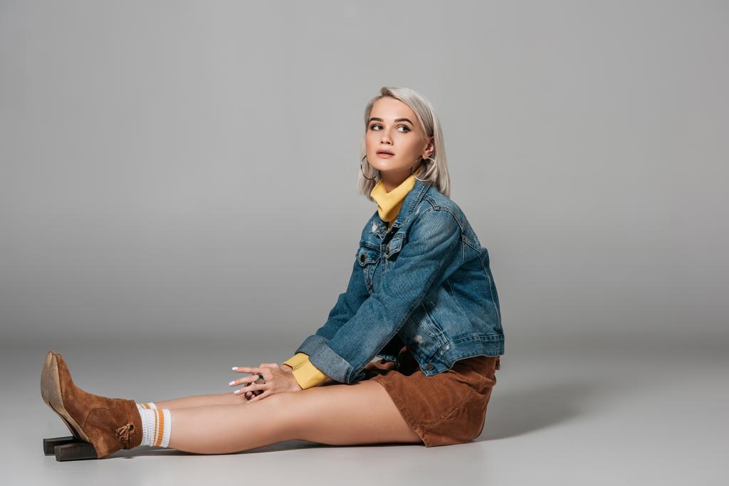 приваблива блондинка модель, що сидить у модній спідниці та джинсовій куртці, на сірому
 - Фото, зображення