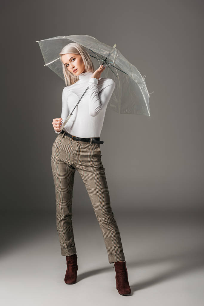 приваблива блондинка в білому черепашку і сірі штани позують з прозорою парасолькою, на сірому
 - Фото, зображення