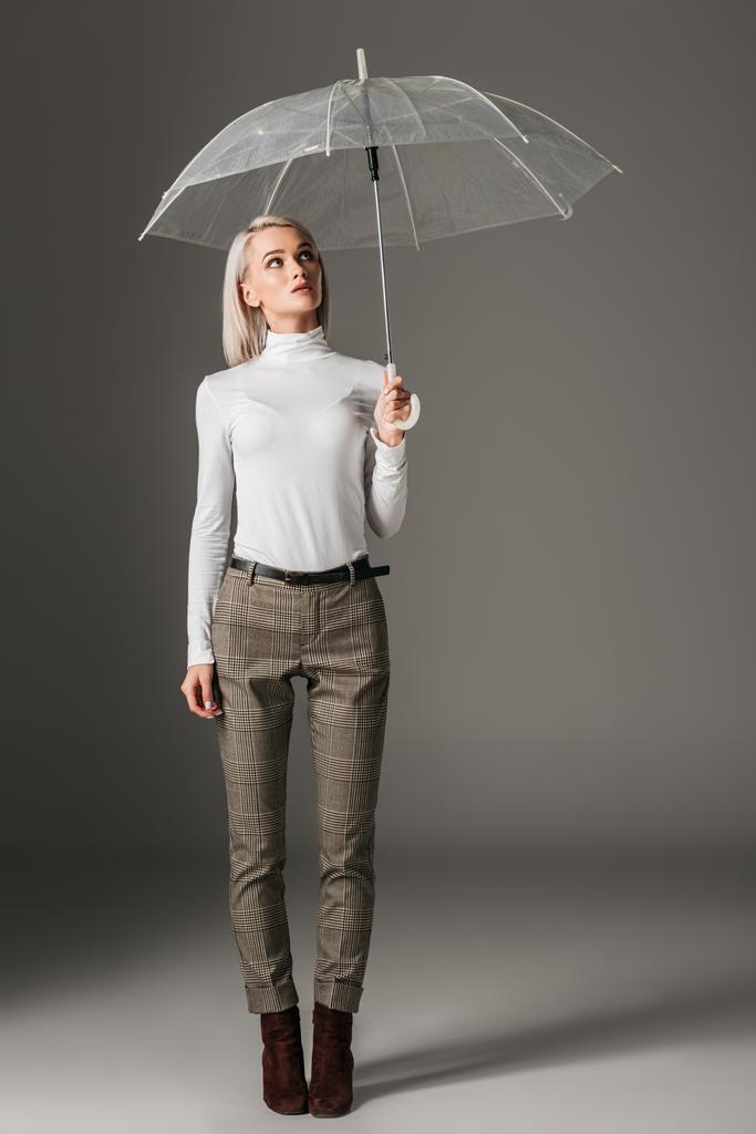 елегантна модна жінка в білому черепашку і сірі штани позують з прозорою парасолькою, на сірому
 - Фото, зображення