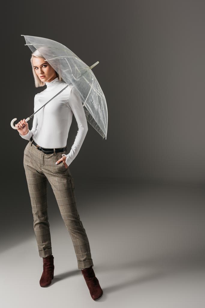 elegante chica con estilo en cuello alto blanco y pantalones grises posando con paraguas transparente, en gris
 - Foto, Imagen