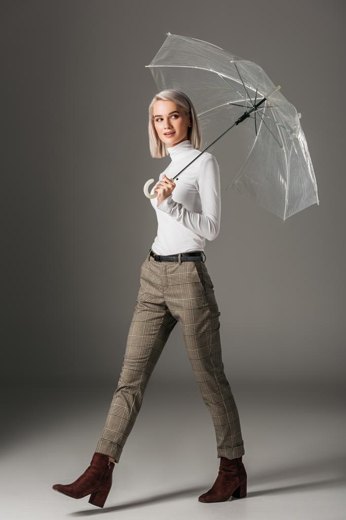 elegant blonde girl in white turtleneck and grey pants walking with transparent umbrella, on grey - Photo, Image