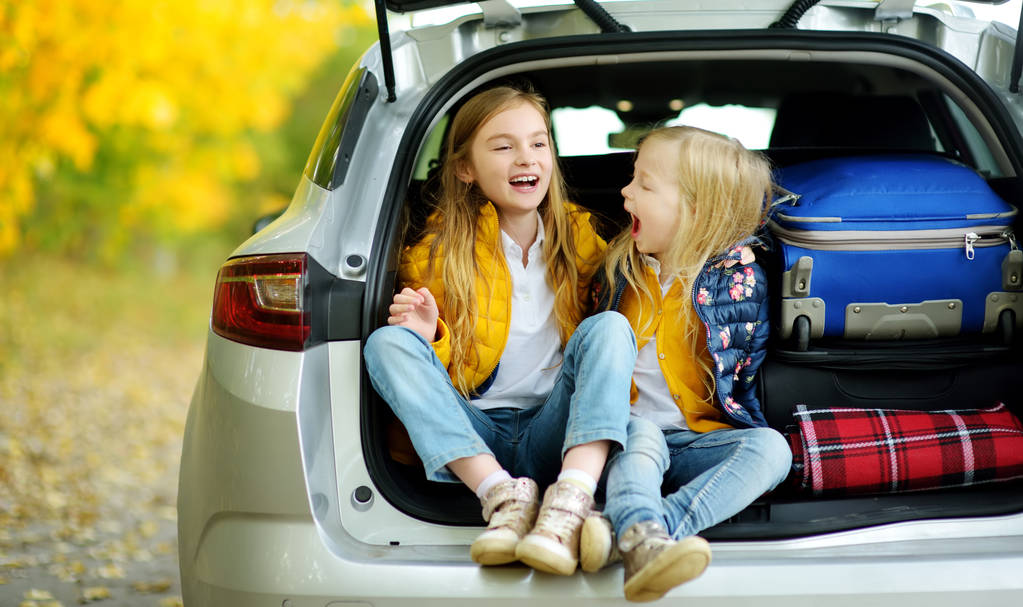 Δύο αξιολάτρευτο κορίτσια που κάθονταν σε ένα κορμό του αυτοκινήτου πριν από τις διακοπές με τους γονείς τους. Δύο παιδιά για ένα οδικό ταξίδι ή ταξίδια με ανυπομονησία. Φθινόπωρο διάλειμμα στο σχολείο. Ταξίδια με το αυτοκίνητο. - Φωτογραφία, εικόνα
