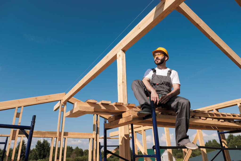 The man builder sits on the edge of the roof of the frame house, in a yellow helmet and gray overalls. The blue sky and clear sunny day. - Photo, Image