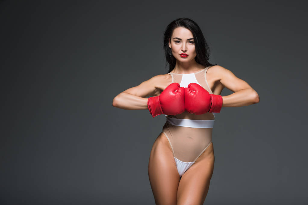 sexy mujer deportiva en traje de baño blanco y guantes de boxeo mirando a la cámara aislada en gris
 - Foto, Imagen
