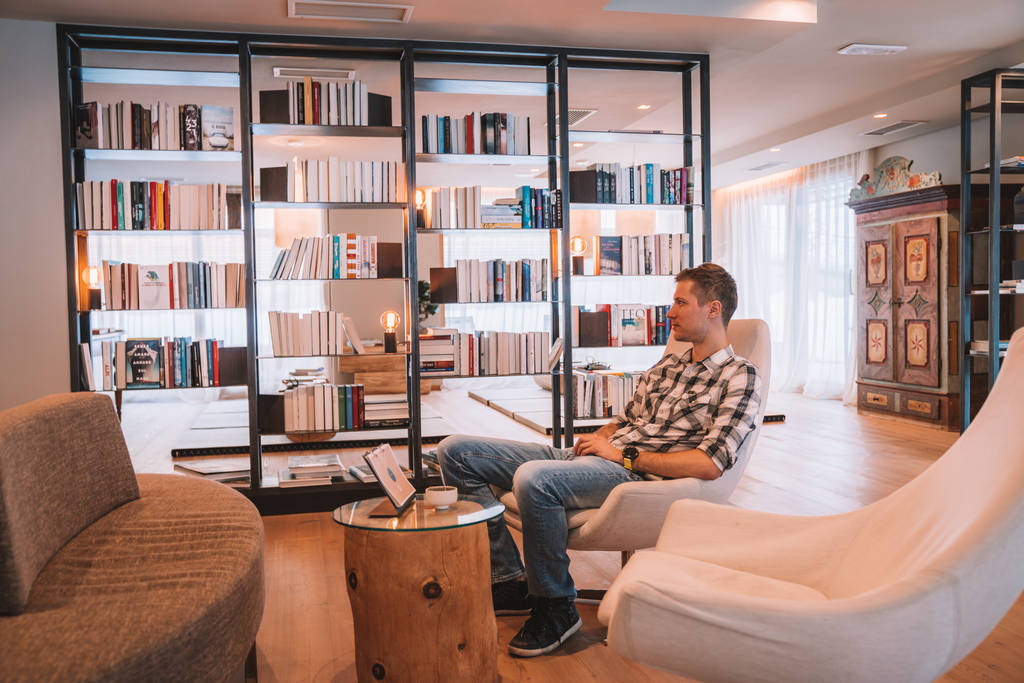 Saalbach, Áustria. Hotel Alpina. 20 de março de 2018. Jovem sentado em uma cadeira em uma confortável sala de biblioteca perto de enormes janelas
. - Foto, Imagem