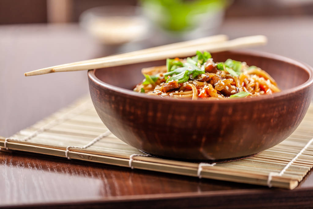 Asian Oriental Cuisine. Japanese noodles with chicken and vegetables, sesame and parsley, in a plate of red clay, stands on a table in a restaurant. Copy space, selective focus - Photo, Image