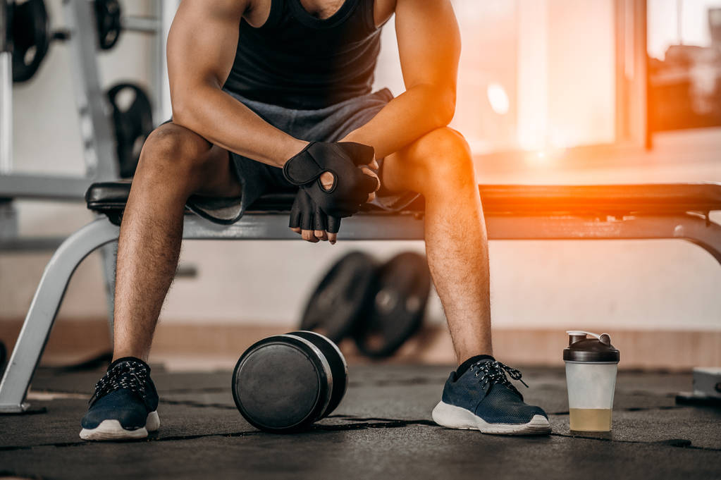 Muskularny mężczyzna na siłowni robi sobie przerwę od treningu siedząc obok butelki soli mineralnej i hantli . - Zdjęcie, obraz