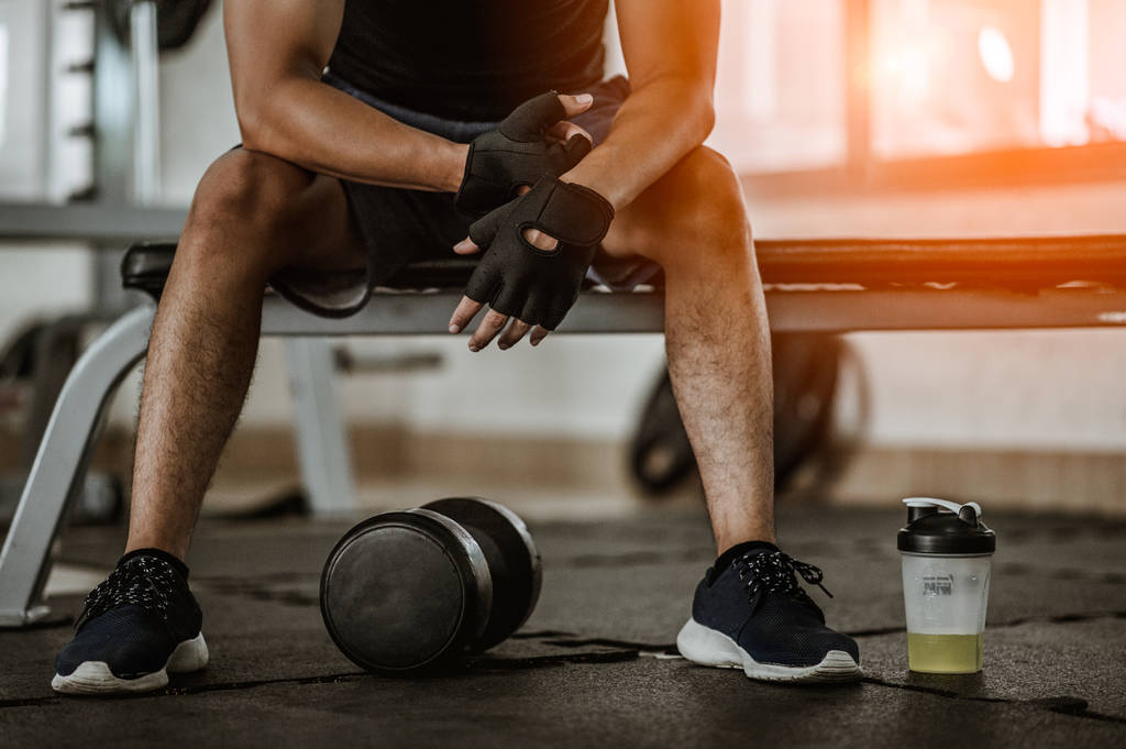 Hombre musculoso en el gimnasio tomando un descanso del entrenamiento sentado al lado de la botella de sal mineral y pesas
 . - Foto, Imagen
