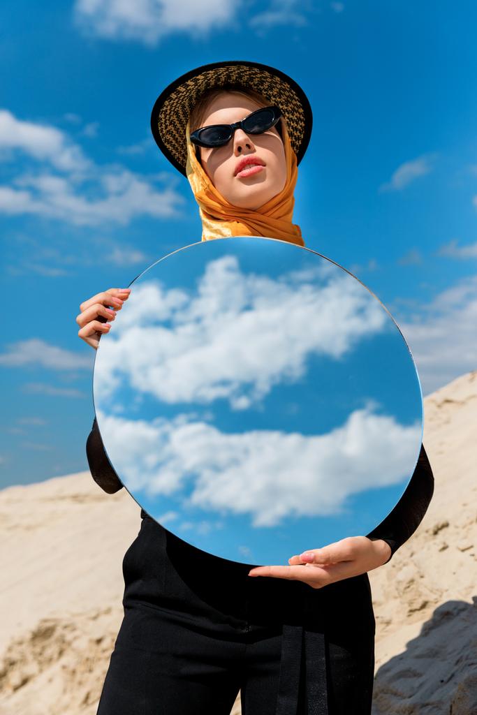 μοντέρνα ελκυστική κοπέλα ποζάρει με στρογγυλό καθρέφτη και αντανάκλαση του συννεφιασμένο ουρανό - Φωτογραφία, εικόνα