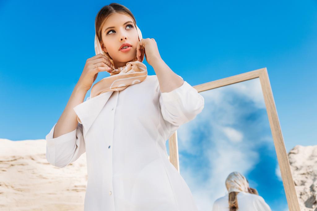 κομψή γυναίκα που θέτουν σε λευκά ρούχα και μεταξωτό μαντήλι κοντά καθρέφτη   - Φωτογραφία, εικόνα