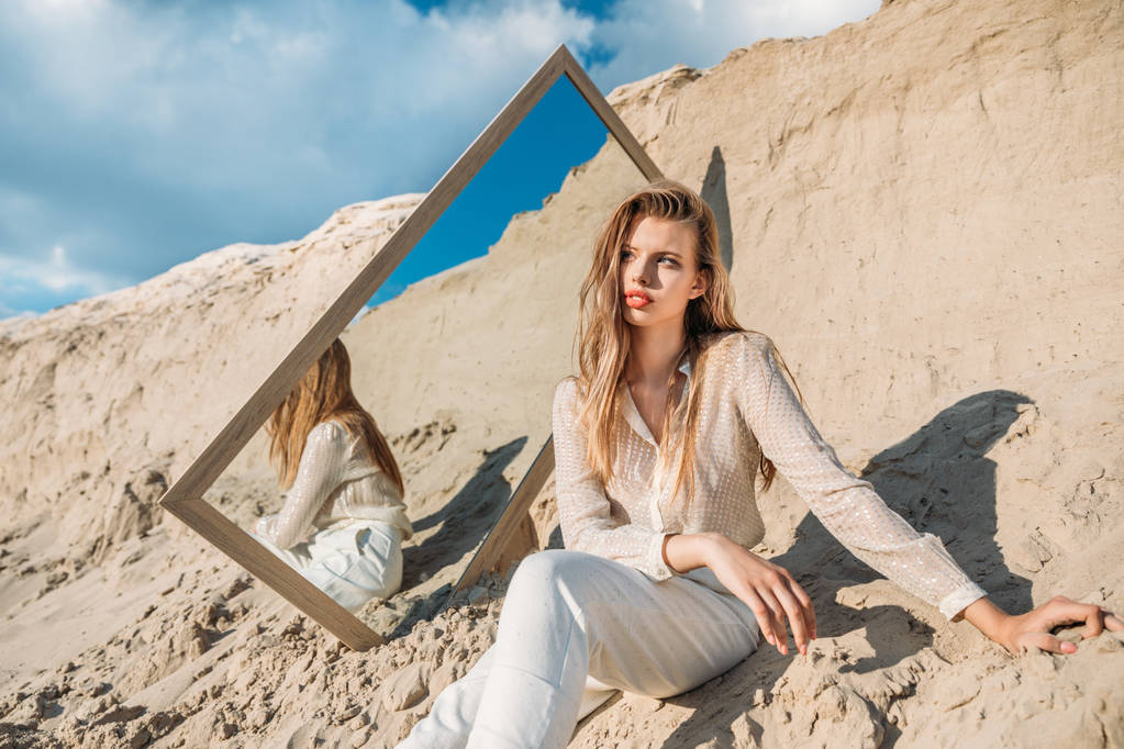 エレガントな若い女性が白い服を着て砂の上の鏡の近くでポーズ - 写真・画像